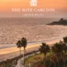 An elevated view of the Laguna Niguel coastline at sunset. A winding coastal path, lined with tall palm trees and green spaces, follows the edge of a sandy beach. The Pacific Ocean is calm, and the sky glows with soft orange and pink colors. The Ritz-Carlton, Laguna Niguel logo is in the bottom right corner.