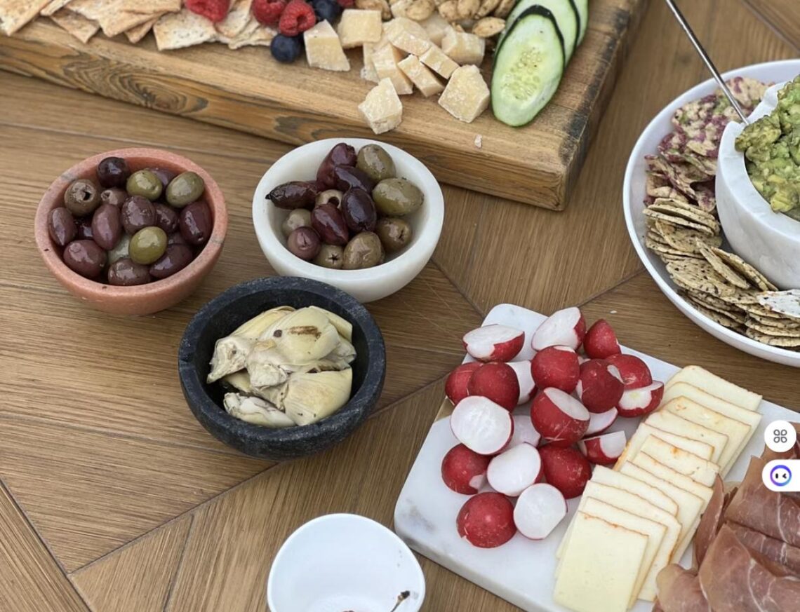 An overhead view of a colorful charcuterie spread with olives, radishes, cheese, crackers, and guacamole on a wooden table.
