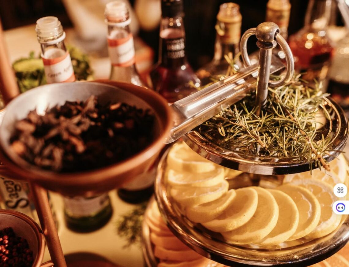 A build-your-own cocktail garnish station with dried citrus, herbs, small mixer bottles, and spice bowls on a bar cart.