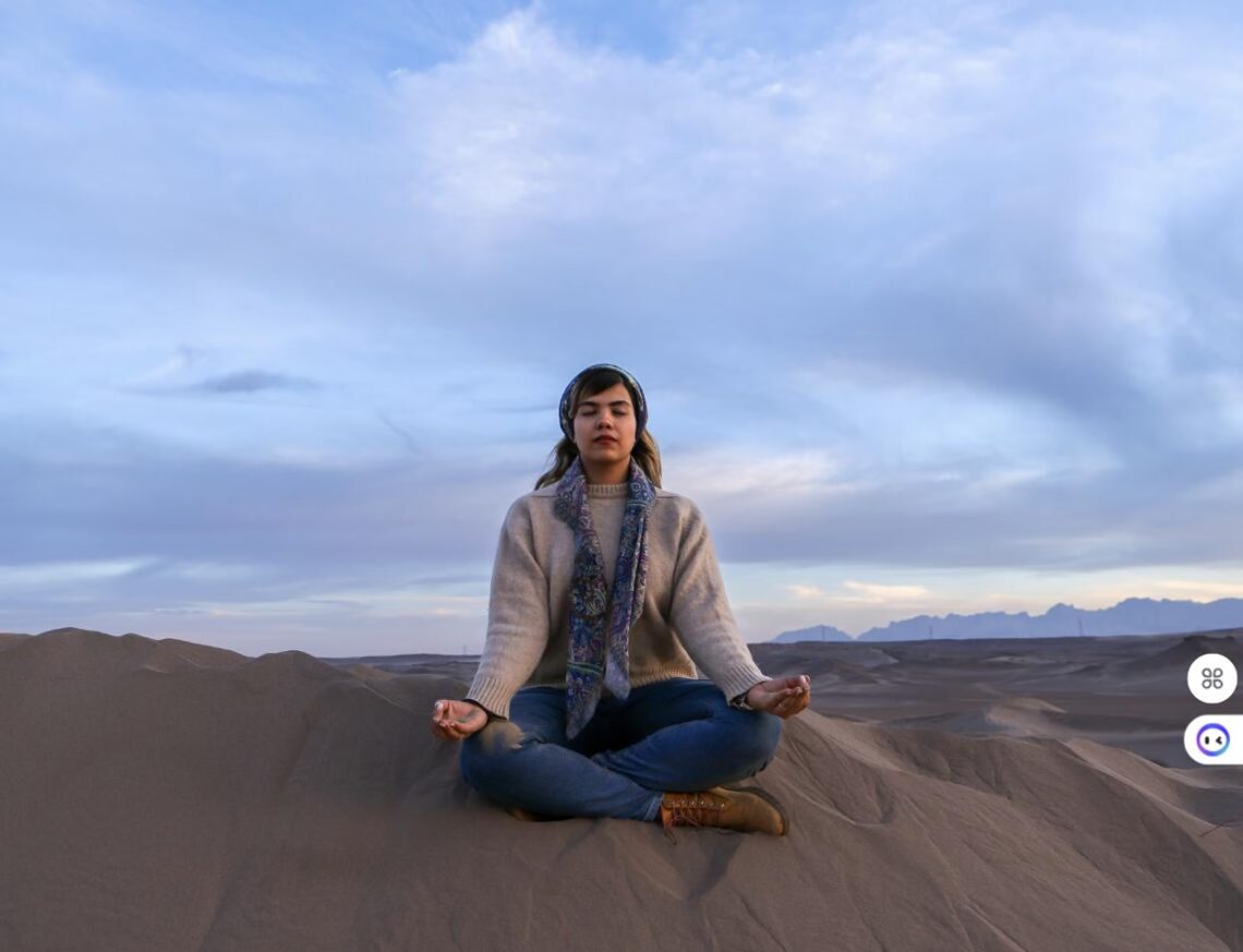 A woman meditates cross-legged on a sand dune in a calm desert landscape at sunset.