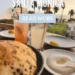 A top-down view of a table with two cocktails, flatbread, and a bowl of hummus, overlooking the Santa Monica coast with palm trees at sunset. White text reads "BEST RESTAURANTS SANTA MONICA" and a light blue button says "READ MORE".