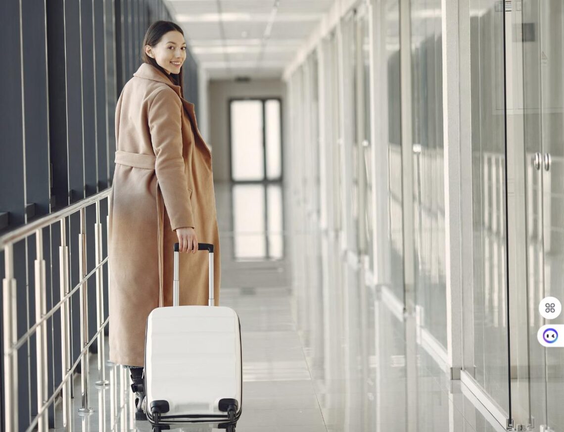 A woman in a long camel-colored coat pulls a white suitcase through a modern airport hallway.