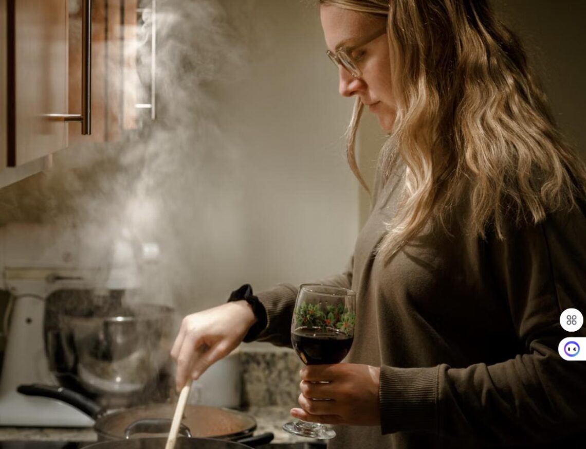 A woman stirs a steaming pot on the stove while holding a glass of red wine in a cozy kitchen.