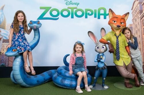 Three happy young girls posing with large Disney Zootopia statues in a movie theater lobby. On the left, Madeline sits on top of a large blue snake statue wearing a blue floral dress. In the center, Charlotte sits on the snake's lower coil wearing a pink shirt, denim overalls, and a blue bow headband. On the right, a friend leans against the Nick Wilde fox statue next to the Judy Hopps rabbit statue.