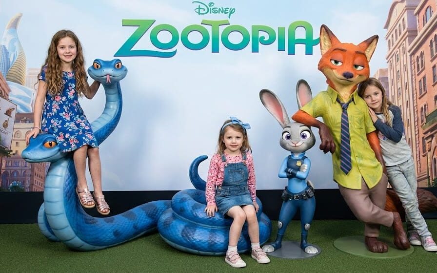 Three happy young girls posing with large Disney Zootopia statues in a movie theater lobby. On the left, Madeline sits on top of a large blue snake statue wearing a blue floral dress. In the center, Charlotte sits on the snake's lower coil wearing a pink shirt, denim overalls, and a blue bow headband. On the right, a friend leans against the Nick Wilde fox statue next to the Judy Hopps rabbit statue.