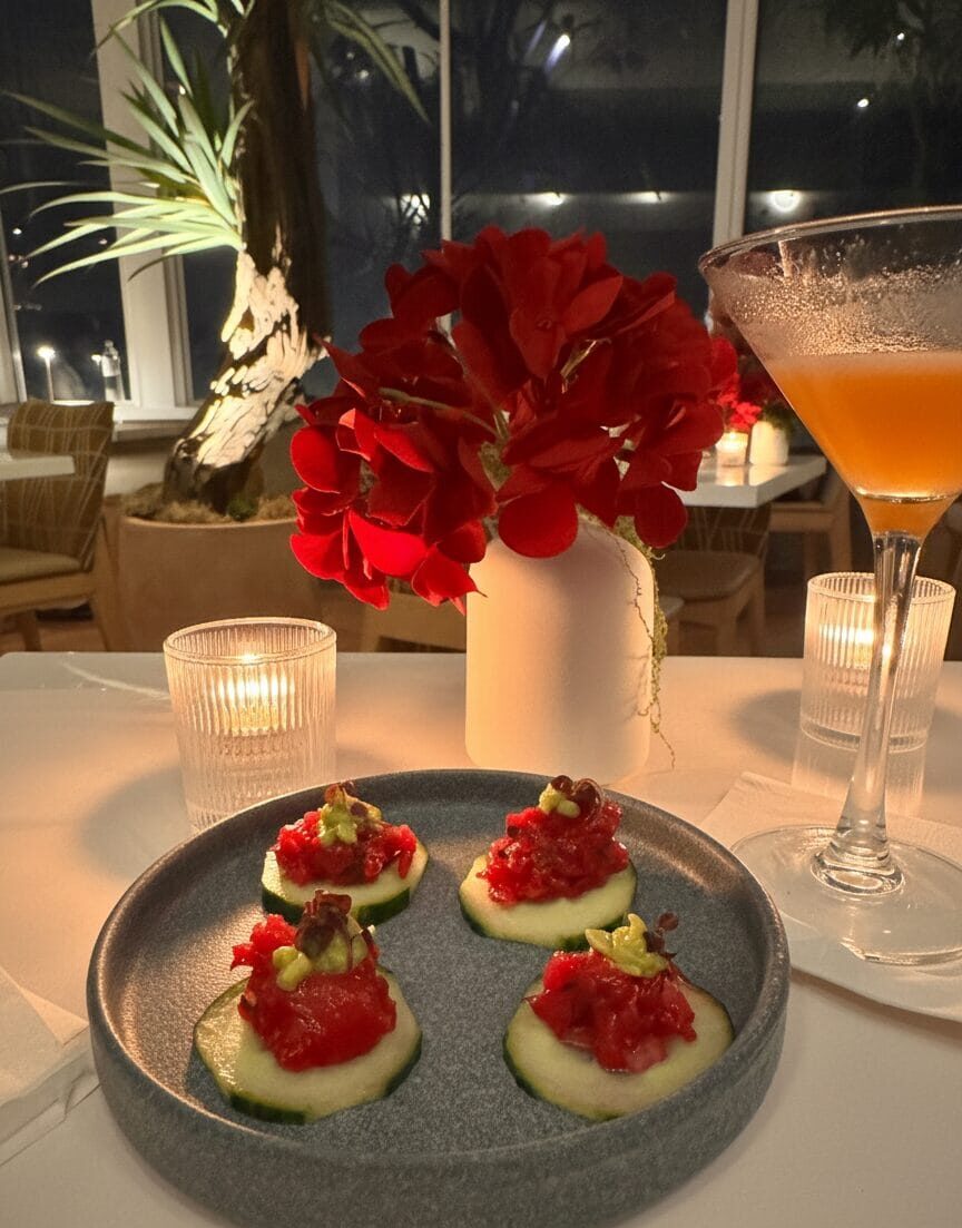Tuna tartare appetizer bites on cucumber slices next to a martini glass with an orange cocktail and a red flower centerpiece in a dimly lit, upscale Santa Monica hotel restaurant.