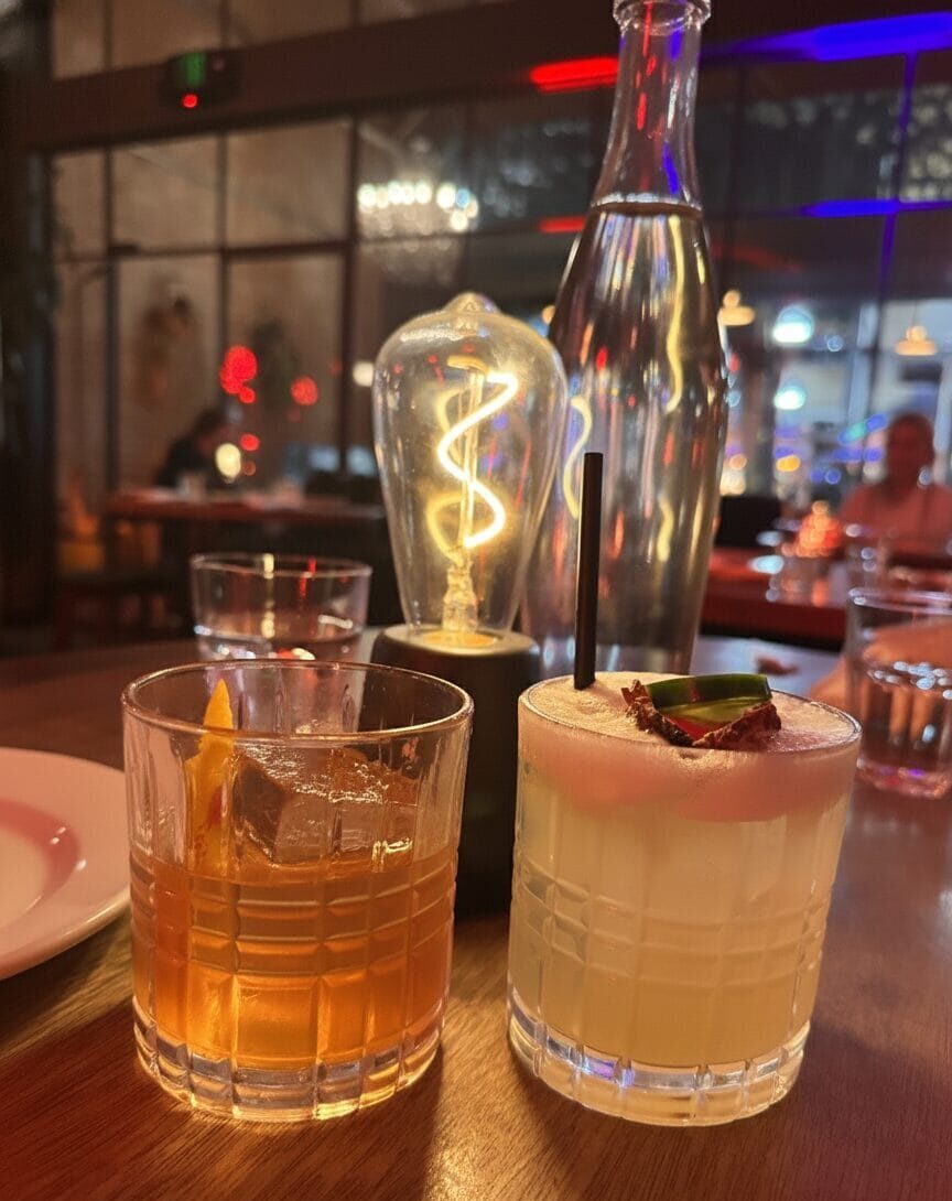 Calvados Old Fashioned and Mezcalita D’Ananas cocktails on a wooden high-top table at Massilia restaurant in Santa Monica, featuring an Edison bulb lamp and a decanter of water in a dimly lit, speakeasy-style setting.