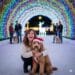 Ginger Graham smiling brightly while kneeling next to her Goldendoodle, Barnaby, who is wearing a red Santa hat and looking directly at the camera. They are posing inside a massive, arched tunnel of colorful Christmas lights, with a white, illuminated reindeer statue in the background. Other people with dogs are visible enjoying the festive light display further down the path.