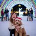 Ginger Graham smiling brightly while kneeling next to her Goldendoodle, Barnaby, who is wearing a red Santa hat and looking directly at the camera. They are posing inside a massive, arched tunnel of colorful Christmas lights, with a white, illuminated reindeer statue in the background. Other people with dogs are visible enjoying the festive light display further down the path.