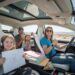 A funny photo of the Graham family (Brad, Ginger, Charlotte, and Madeline) on their LA to Vegas road trip in a Tesla. Ginger is driving with a confident thumbs-up and sunglasses, while Brad in the back seat looks bewildered and is eating a snack. The two daughters, Charlotte and Madeline, are happy Madeline is proudly holding up a drawing, and Charlotte is eating a snack. The car is packed with books, snacks, and gear.