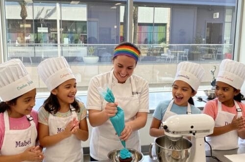 Chef and five giggling 8-year-old girls in chef hats and personalized aprons baking cookies and cupcakes in a professional kitchen at The Gourmandise School in Santa Monica.