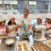 Chef and five giggling 8-year-old girls in chef hats and personalized aprons baking cookies and cupcakes in a professional kitchen at The Gourmandise School in Santa Monica.