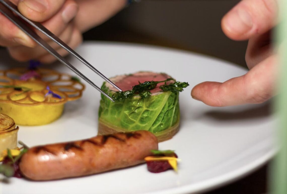A chef uses tweezers to carefully plate a gourmet dish with meat, sautéed greens, and grilled sausage on a white plate.