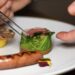 A chef uses tweezers to carefully plate a gourmet dish with meat, sautéed greens, and grilled sausage on a white plate.