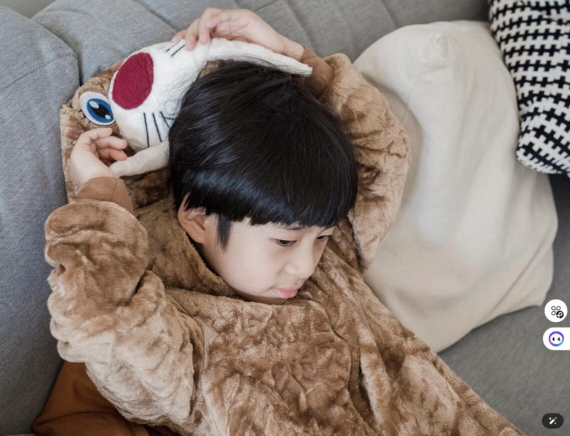 A child cuddles a stuffed animal while wrapped in a cozy plush wearable blanket on the couch.