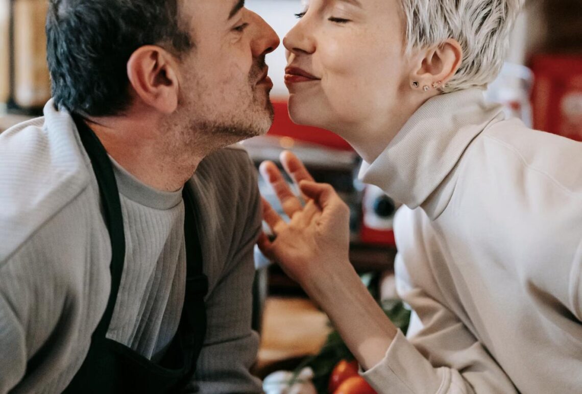 A couple leans in close while cooking together in the kitchen, sharing a playful, romantic moment.