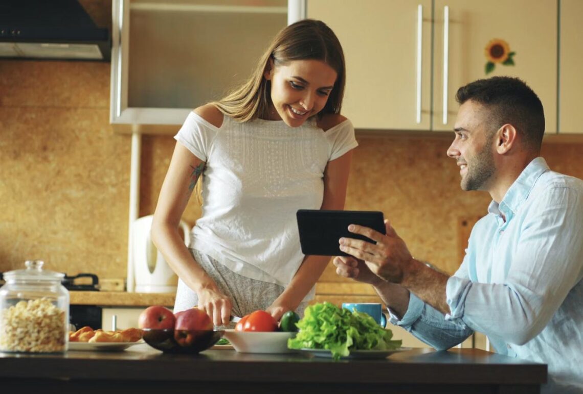 A couple looks at a tablet together while preparing vegetables in the kitchen, planning their meal.