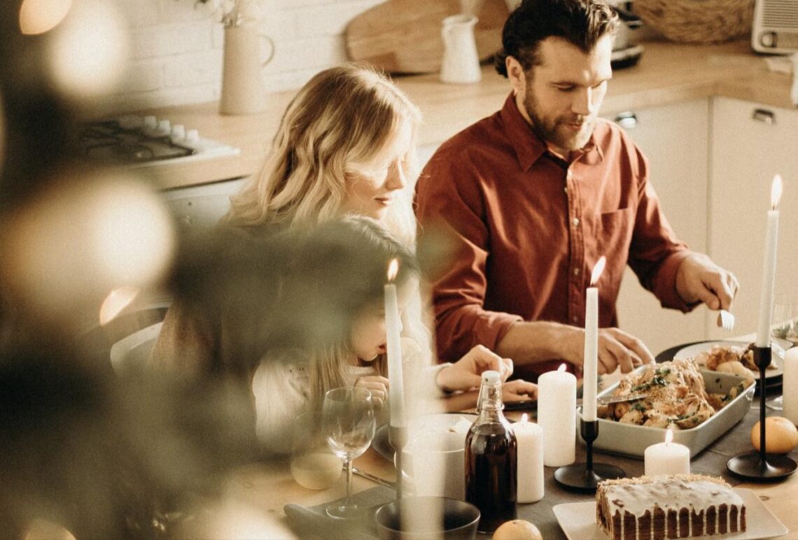 A family gathers in the kitchen preparing a holiday meal together, with a young girl looking on as a man carves a roast at a candlelit table.