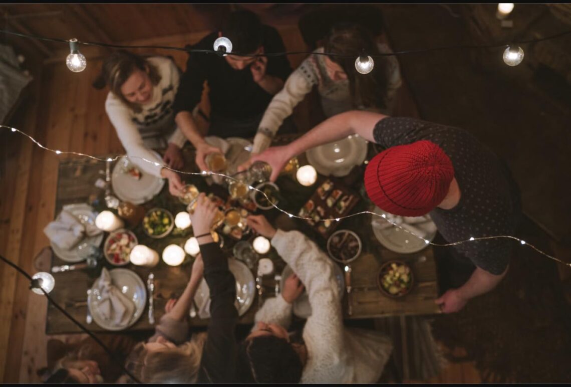 Overhead view of a group of friends reaching across a candlelit table to share dishes at a festive dinner party.