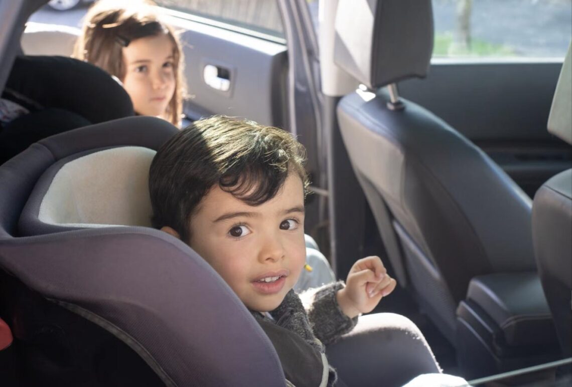 Two young children buckled into car seats in the back seat, looking toward the camera during a road trip.