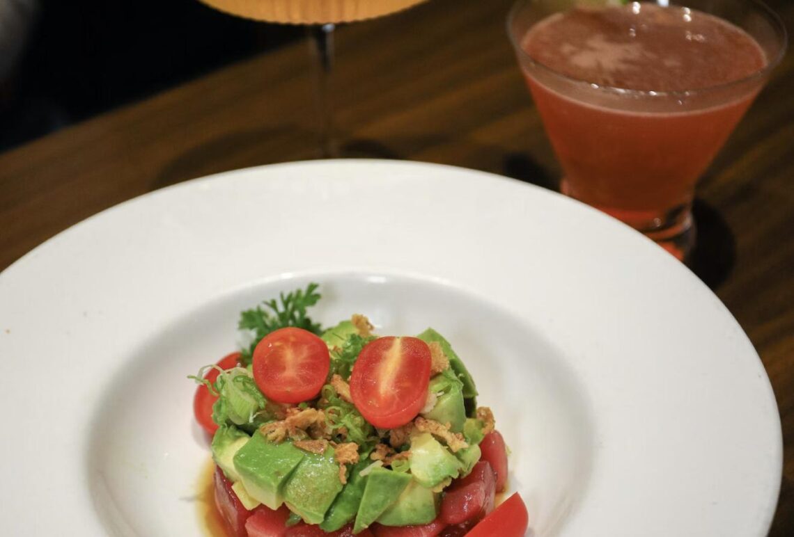 A plate of tuna tartare with avocado and tomato beside a red cocktail on a restaurant table.