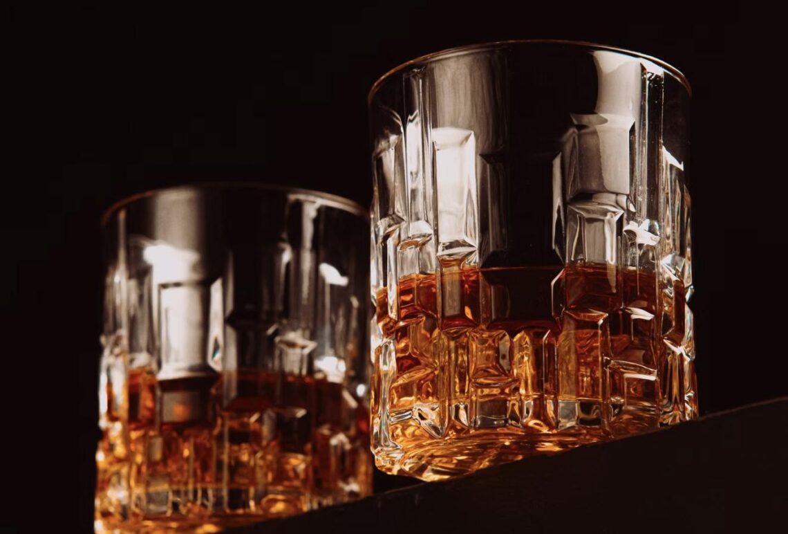 Two whiskey cocktails on the rocks, glowing amber against a dark, moody bar background.