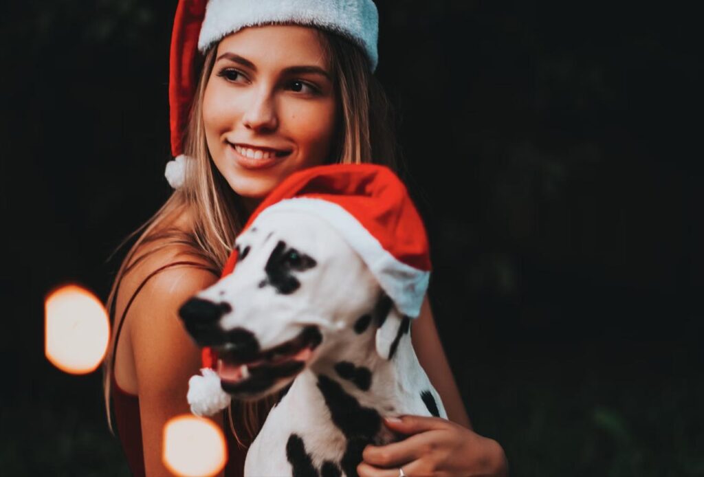 A smiling woman wearing a Santa hat holds her dog, who is also wearing a Santa hat, at a nighttime holiday light display.