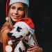 A smiling woman wearing a Santa hat holds her dog, who is also wearing a Santa hat, at a nighttime holiday light display.