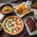 A top-down view of a wooden table featuring a bowl of Jinya ramen, a HiHo cheeseburger with fries, a Pizzana margherita and spinach pizza, and a tray of Wood Ranch BBQ ribs.