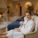 A woman in a white luxury bathrobe relaxing in a plush spa lounge with a warm drink, representing a wellness retreat at Canyon Ranch Spa in Las Vegas.