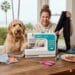 A blonde woman and her Goldendoodle dog sit at a wooden table with a Brother CS7000X sewing machine. She is laughing and holding up a pair of poor quality black leggings with a torn seam, while the dog has a paw on the machine as if helping. A plate of fresh tuna crudo sits on the table nearby.