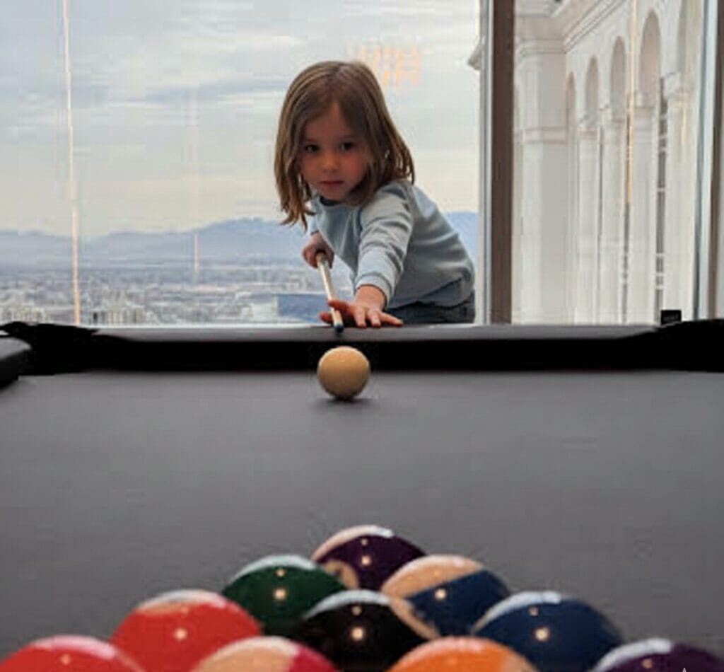 A first-person perspective of a pool table with a rack of billiard balls in the foreground and a single cue ball centered in the distance, set against a floor-to-ceiling window overlooking a sprawling city and mountain range.