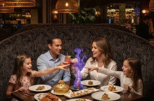 A family of four sits in a dark, patterned booth at an upscale restaurant, smiling as they watch a blue flambé fire rise from a dish on their table while the parents toast with pink cocktails.