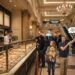 A woman holding a Goldendoodle puppy standing in a long line at Bouchon Bakery in the Venetian Hotel, while her husband points urgently toward an escalator leading to a Coffee Bean & Tea Leaf sign.