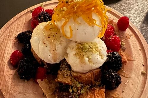 A luxury dessert featuring three large scoops of frozen greek yogurt stacked on crisp phyllo baklava, topped with 24k gold leaf honey and candied citrus garnished with fresh raspberries and blackberries on a round wooden board.