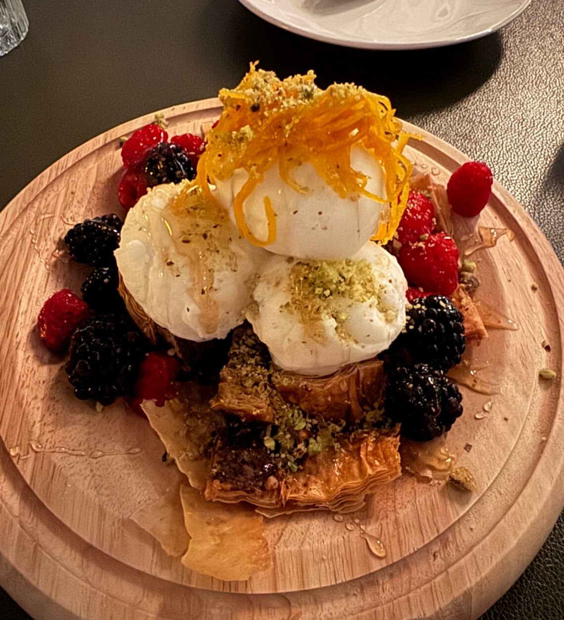 A luxury dessert featuring three large scoops of frozen greek yogurt stacked on crisp phyllo baklava, topped with 24k gold leaf honey and candied citrus garnished with fresh raspberries and blackberries on a round wooden board.