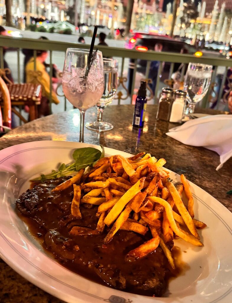 A plate of steak frites on a restaurant patio table at night with a view of the Bellagio fountains and hotel in Las Vegas.