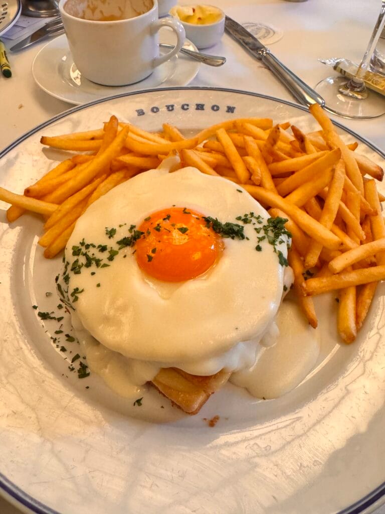 A gourmet Croque Madame topped with a fried egg and Mornay sauce, served with golden French fries on a signature Bouchon Bakery plate next to a cup of espresso.