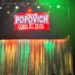 A view from the audience of the Popovich Comedy Pet Theater stage in Las Vegas, featuring a bright red neon sign and gold curtains under colorful stage lights.