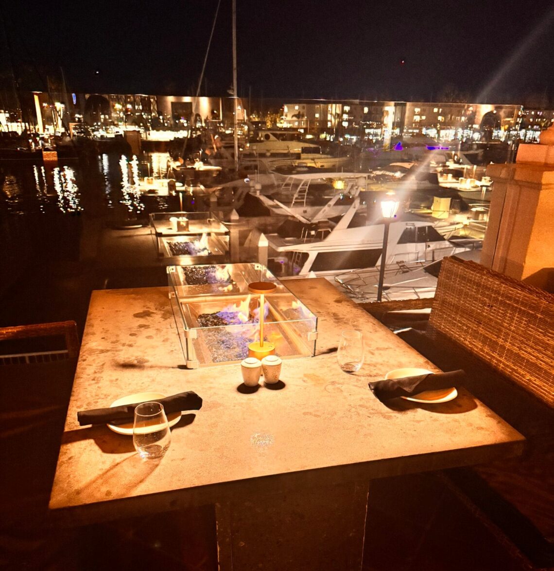 Outdoor fire-pit dining table at Cast & Plow in the Ritz-Carlton Marina del Rey overlooking luxury yachts at night.