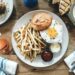 An overhead view of a burger and fries with a fried egg on top, served alongside a cocktail and other dishes on a wooden table.