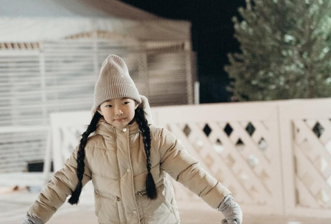 A young girl in a winter coat and beanie stands at an outdoor ice rink lit with string lights at night.