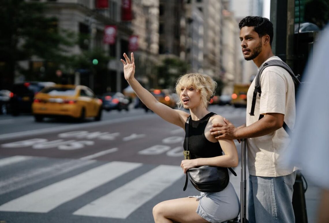 A woman hails a taxi on a busy city street while her travel companion waits beside their luggage.