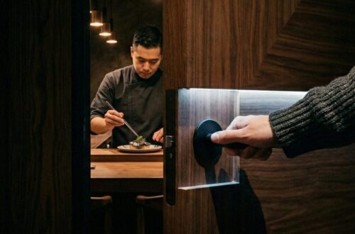 A hand in a charcoal ribbed knit sleeve opening a secret walnut door to reveal Chef Brian Baik preparing a dish at the Corridor 109 seafood counter in Melrose Hill, Los Angeles. High-contrast cinematic lighting.
