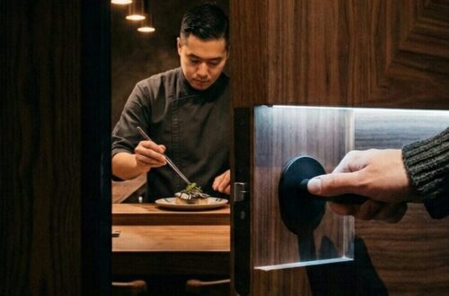 A hand in a charcoal ribbed knit sleeve opening a secret walnut door to reveal Chef Brian Baik preparing a dish at the Corridor 109 seafood counter in Melrose Hill, Los Angeles. High-contrast cinematic lighting.