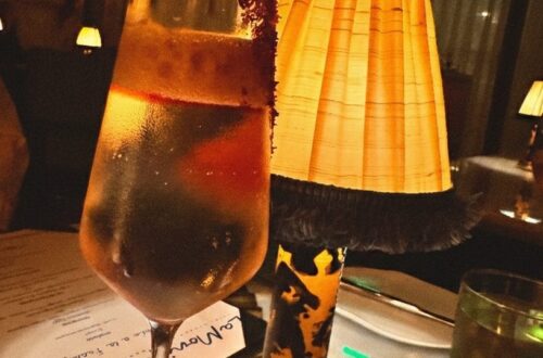 A sophisticated dining scene at La Monique in Santa Monica featuring a clarified cocktail with a garnet rim next to a vintage-style pleated lampshade and a prix-fixe Valentine's Day menu on a white tablecloth.