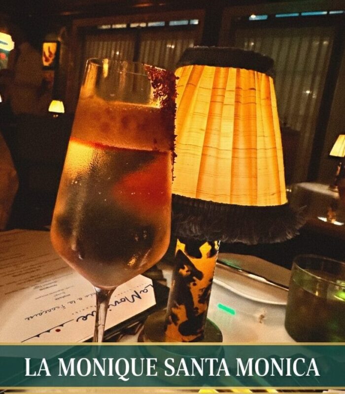 A sophisticated dining scene at La Monique in Santa Monica featuring a clarified cocktail with a garnet rim next to a vintage-style pleated lampshade and a prix-fixe Valentine's Day menu on a white tablecloth.