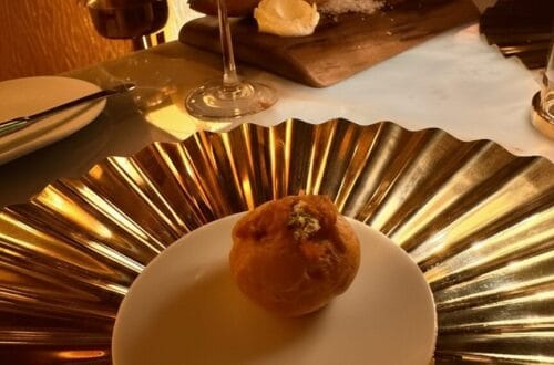 A close-up of a delicate amuse-bouche served on a small white plate, resting atop a large, reflective gold-pleated decorative charger. In the background, a chilled glass of rosé wine and a rustic bread service on a wooden board are illuminated by warm, moody restaurant lighting.