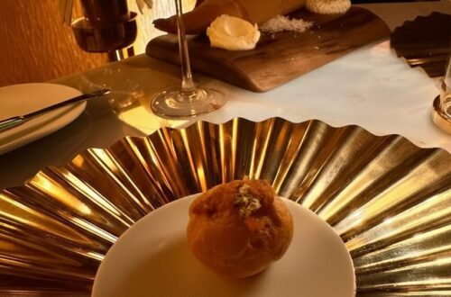 A close-up of a delicate amuse-bouche served on a small white plate, resting atop a large, reflective gold-pleated decorative charger. In the background, a chilled glass of rosé wine and a rustic bread service on a wooden board are illuminated by warm, moody restaurant lighting.