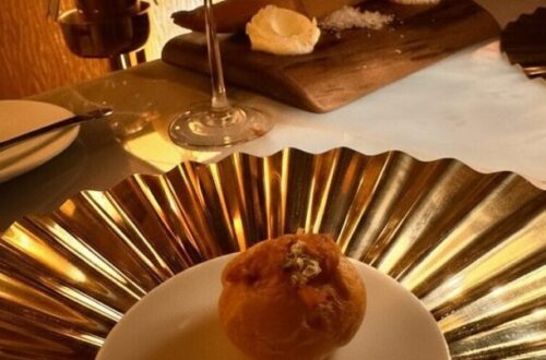 A close-up of a delicate amuse-bouche served on a small white plate, resting atop a large, reflective gold-pleated decorative charger. In the background, a chilled glass of rosé wine and a rustic bread service on a wooden board are illuminated by warm, moody restaurant lighting.